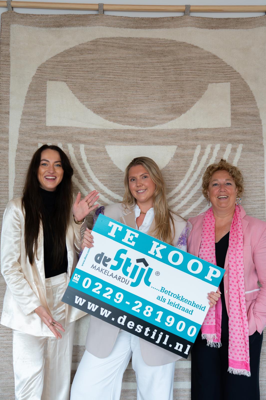 Drie vrouwelijke teamleden van De Stijl Makelaardij houden samen een ‘Te Koop’-bord vast, met de slogan ‘Betrokkenheid als leidraad’.