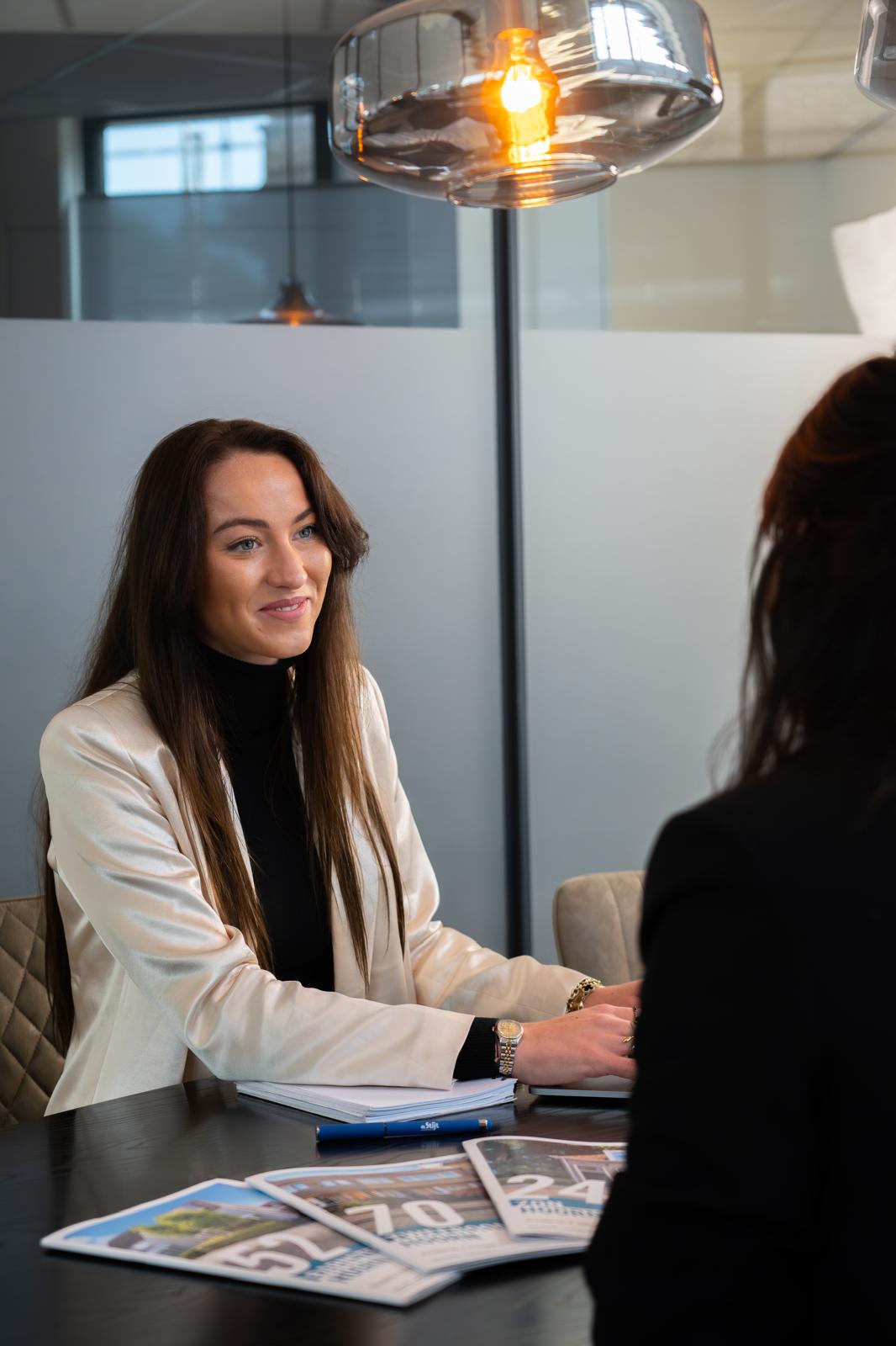 Een makelaar van De Stijl Makelaardij zit aan tafel in gesprek met een klant, met brochures van woningen op de voorgrond.